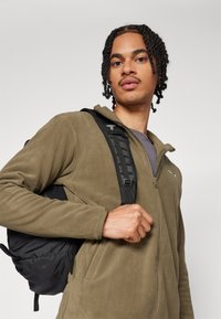 Olive green fleece zip-up jacket, featuring a textured surface, paired with a black backpack showing adjustable straps and buckle detail.