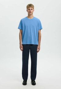 Zils kokvilnas T-krekls ar apaļu kakla izgriezumu, apvienots ar tumši ziliem taisnajiem biksēm un melniem apaviem, modelis stāv pretī vienkāršai fona.