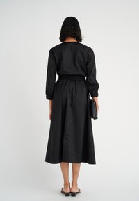 Schwarzes Midi-Kleid mit langen Ärmeln und elastischem Taillengummi, das eine glatte Textur aufweist. Das Model hält eine schwarze gesteppte Handtasche.