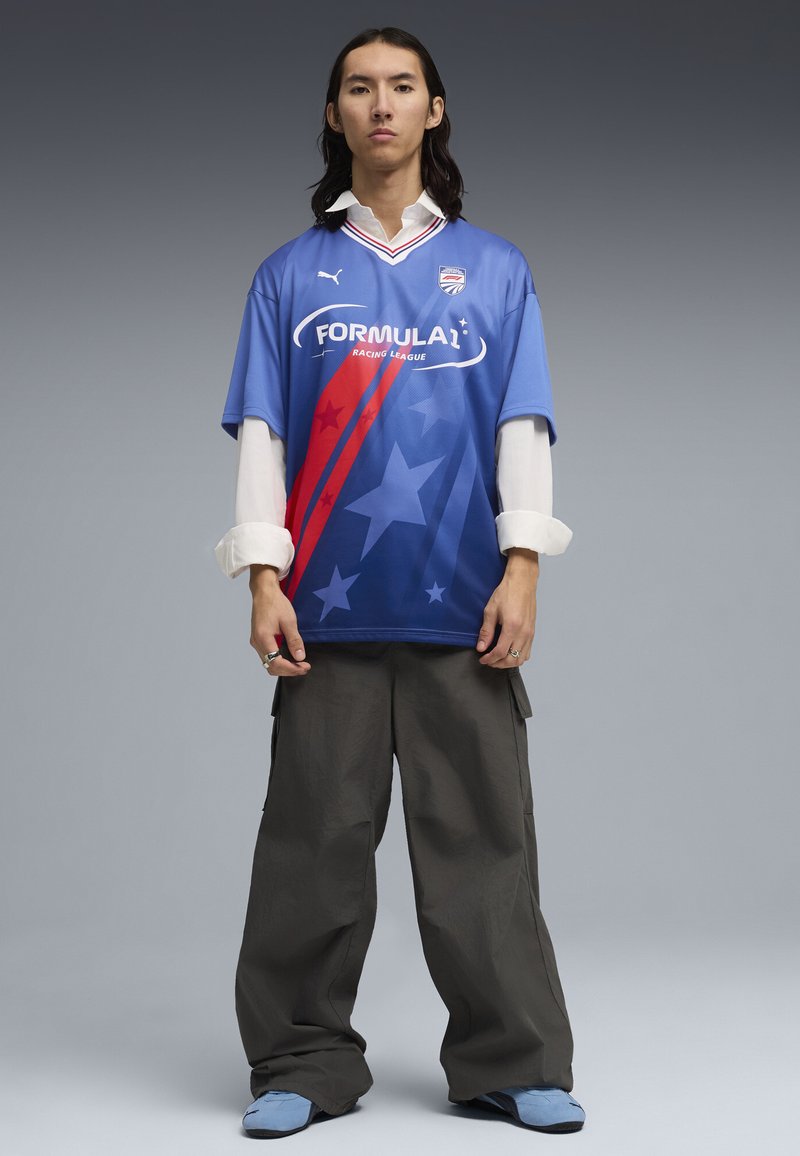 Jeune personne aux longs cheveux portant un t-shirt bleu de la Formula 1 Racing League sur un t-shirt blanc, un pantalon large foncé et des chaussures bleu clair.