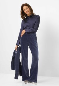 Marineblauw satijnen overhemd met lange mouwen, gecombineerd met marineblauwe corduroy wijde broek. Het model houdt een gestructureerde marineblauwe trui vast. Zilveren schoenen.
