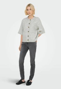 Femme avec des cheveux blonds courts portant un cardigan boutonné gris clair, un jean slim gris foncé et des mocassins noirs sans lacets avec des embellissements en argent.