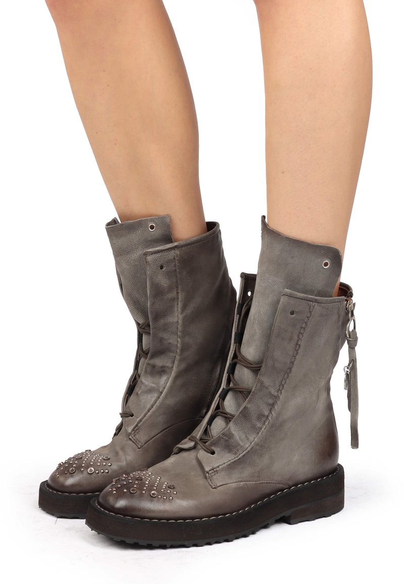 Bottes montantes vintage en cuir gris avec lacets, détails cloutés sur les orteils, portées sur des jambes nues, avec des tirettes à glissière latérales.