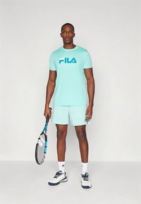 Ein mintgrünes Sportoutfit mit einem Fila-Logo-T-Shirt und passender Shorts, kombiniert mit einem Tennisschläger und weißen Sneakern.