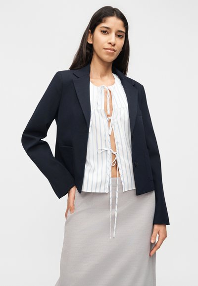 Γυναίκα που φοράει μαύρο σακάκι πάνω από λευκό μπλούζα με κάθετες μπλε ρίγες δεμένη με τρία φιόγκους, και ανοιχτό γκρι φούστα, στέκεται μπροστά σε λευκό φόντο.