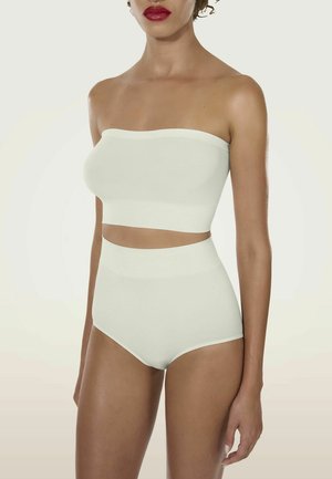 Donna snella che indossa un bandeau bianco senza cuciture e senza spalline e slip a vita alta, in piedi davanti a uno sfondo chiaro e uniforme.