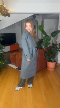 Manteau long gris avec un design portefeuille, doté d'une ceinture et de poignets boutonnés. Porté avec un pantalon vert et assorti à des baskets grises. Fond en parquet en bois.
