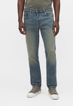 Homme portant un jean bleu délavé droit, un t-shirt vert olive et des baskets basses grises, debout sur un fond blanc.