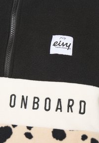 Veste en polaire avec une partie supérieure noire, une bande crème portant le texte "ONBOARD", et une partie inférieure beige avec des taches noires. Détail de fermeture éclair visible.