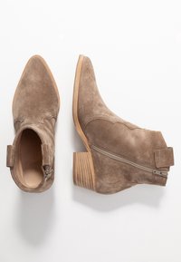 Botas de tobillo de ante marrón con punteras puntiagudas, tacones de madera apilada, cremalleras laterales y costuras decorativas en la parte superior.