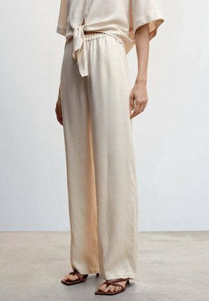 Vrouw draagt losse lichtbeige linnen broek in combinatie met bijpassende top met strik aan de voorkant en bruine platte sandalen met bandjes op een betonnen vloer.