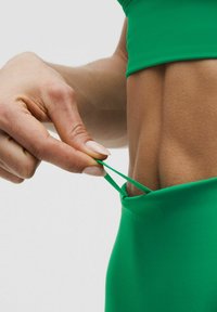 Žalios sportinės legging kelnės su lygiu, elastingu paviršiumi; ranka traukia raištelį iš juosmens, išryškindama jų pritaikymą ir dizainą.