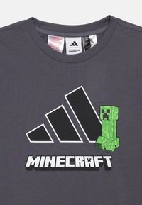 Šedé tričko s výraznou zelenou pixelovanou grafikou Creepera z Minecraftu, s bielym logom Adidas a textom "MINECRAFT" v blokovom štýle.