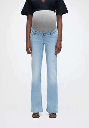 Nosečnica v svetlo modrih nosečniških kavbojkah s sivim trebušnim delom, črni majici in črnih sandalih z odprtimi prsti stoji z rokami sproščenimi ob telesu.