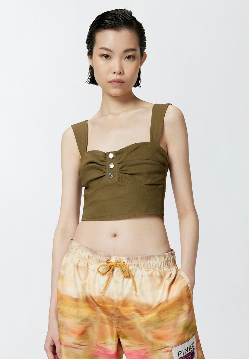 Top cropped verde oliva con spalline larghe, fronte arricciato e dettagli con bottoni. La modella indossa pantaloni corti a fantasia colorata con cordoncino.