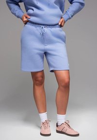 Shorts in cotone azzurro chiaro con vita elasticizzata e cordino. Presenta un piccolo logo sulla gamba. Indossati con scarpe da ginnastica rosa e calzini bianchi.