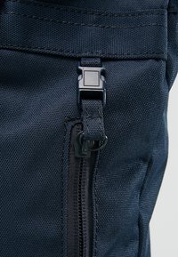 Tmavě modrá textilní taška s texturovaným povrchem, plastovou sponou a zipem podél boku pro uzavření a přístup.