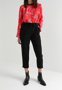 Rote Bluse mit Blumenmuster, langen Ärmeln, gerafftem Ausschnitt und schwarze, taillierte Hose mit einem Gürtel. Schwarze Knöchelstiefel runden das Outfit ab.