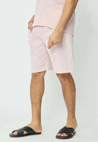 Shorts en lin rose clair avec poches latérales, associés à des sandales noires à lanières croisées. Texture lisse avec une coupe décontractée, longueur mi-cuisse.