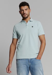 Lichtblauw poloshirt van zachte katoen, met een drieknoopsluiting en een klein hexagonaal logo op de linkerkant van de borst. Korte mouwen.