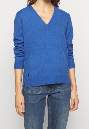 Vrouw die een blauw V-hals trui met lange mouwen en donkerblauwe spijkerbroek draagt, staand tegen een lichte achtergrond.