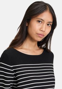 Schwarzer und weißer gestreifter Pullover mit weitem Halsausschnitt, gerippter Textur und langen Ärmeln. Hauptfarbe ist schwarz; die Streifen sind weiß.