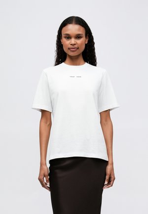 Mujer con cabello rizado largo, vistiendo una camiseta blanca de manga corta con texto pequeño en el pecho y una falda negra, de pie frente a un fondo plano.
