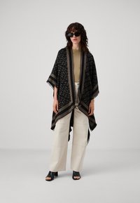 Zwarte en gouden poncho met franjes, voorzien van een herhalend logo-patroon. Gecombineerd met crèmekleurige broek met wijde pijpen en zwarte hoge hakken sandalen.