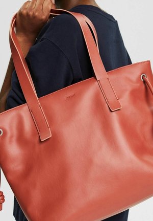 Person trägt ein dunkles Hemd und hält eine große, glatte braune Ledertasche mit doppelten Schulterriemen und dezent geprägt eingraviertem Markennamen.