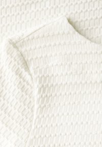 Tessuto bianco testurizzato con un sottile motivo a rombo, caratterizzato da un semplice scollo rotondo e maniche corte. Materiale morbido e leggero.