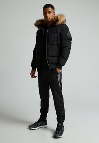 Giubbotto a vento nero con cappuccio bordato in pelliccia, chiusura con zip e patch con logo. Indossato con pantaloni sportivi neri e scarpe da corsa nere.