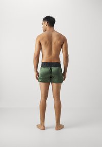 Calções de banho em satin verde com uma cintura elástica preta, apresentando uma textura suave e um corte simples. Usados por uma pessoa vista de trás.