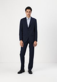 Marineblauer Anzug mit tailliertem Schnitt, ausgestattet mit einer Zwei-Knopf-Verschluss und Reverskragen, kombiniert mit einem hellblauen Hemd und schwarzen Schuhen.