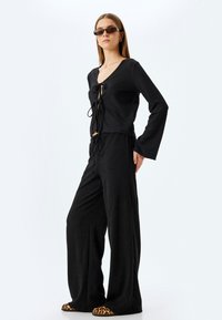 Schwarzes Crop-Top mit V-Ausschnitt und Knoten vorne, kombiniert mit schwarzen weiten Hosen. Die Schuhe haben ein Leopardenmuster als Akzent. Glatte Textur.