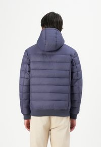 Man wearing a navy blue quilted hooded jacket and beige pants, shown from the back against a plain white background.
