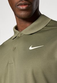 Olívovo zelená polo košeľa vyrobená z textúrovanej látky, s dvojitým gombíkom a bielym logom Nike swoosh na ľavej strane hrude.