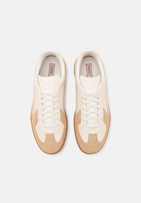 Weiße und beige Sneaker mit Obermaterial aus Wildleder und Leder, flachen Schnürsenkeln, Gummisohle und dezentem Branding an Zunge und Seite.