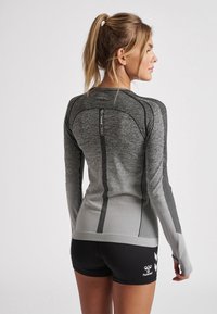 Kvinna med blont hästsvans, klädd i en åtsittande grå långärmad sporttopp och svarta shorts, stående i profil mot en enkel bakgrund.