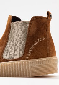 Stivale alla caviglia in suede marrone con un pannello laterale elastico beige testurizzato e una suola in gomma testurizzata con scanalature orizzontali.