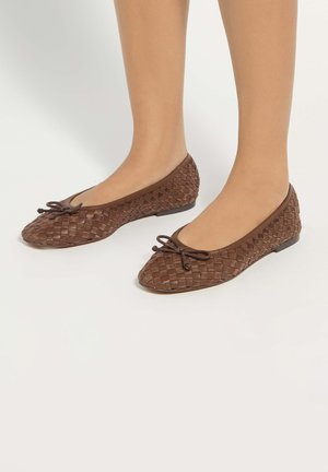Brown woven leather ballet flats with small bow detail worn on bare feet against a plain light background.