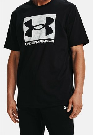 Man draagt een zwarte Under Armour t-shirt met groot logo en tekst op de borst en een bijpassende zwarte broek met klein logo bij de zak.