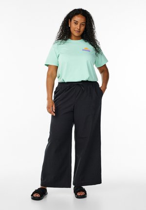 Mintgrünes Kurzarm-T-Shirt mit einem mehrfarbigen Sonnenmotiv; kombiniert mit schwarzen weiten Hosen und schwarzen Rutsch-Sandalen. Einfaches Design.