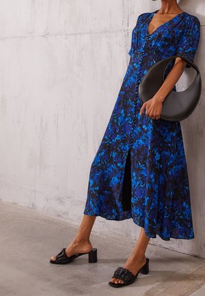 Vrouw draagt een blauw-zwarte bloemen midi-jurk, zwarte sandalen met hak en houdt een zwarte halvemaanvormige handtas vast.