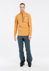 Langärmliger orangefarbener Mid-Layer-Pullover mit einem Reißverschluss-Brusttasche, grauen Akzenten, kombiniert mit dunkelblauen Hosen und schwarzen Skischuhen.