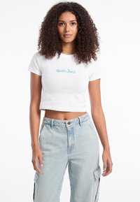 Bjela skraćena majica s turkiznim "Guess Jeans" logotipom, u kombinaciji s svijetloplavim trapericama visokog struka, koje imaju džepove i opušteni kroj.