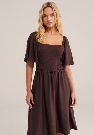 Szőke nő barna, texturált midi ruhában, négyzet alakú nyakkivágással és fodros ujjakkal, egyszínű háttér előtt állva.