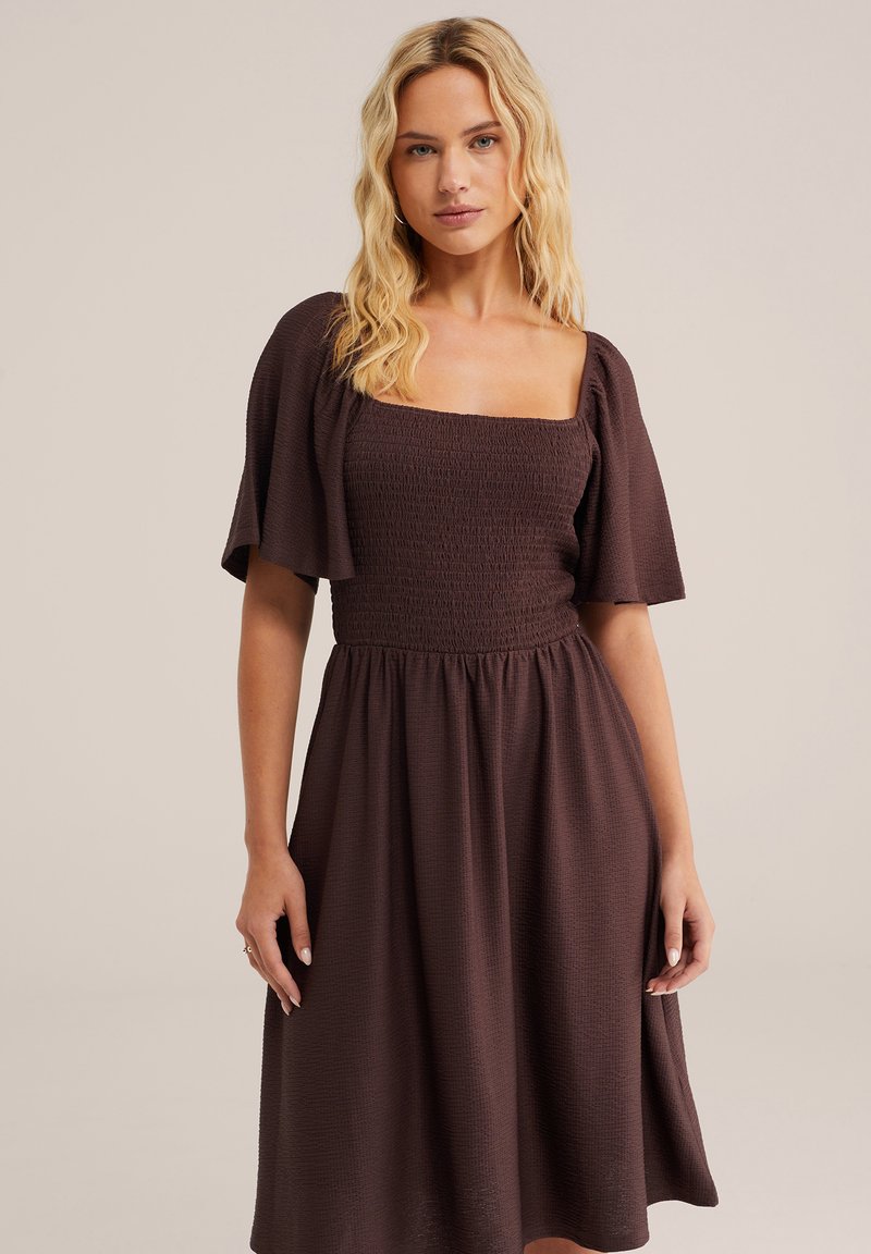 Mujer rubia con un vestido midi marrón texturizado, con cuello cuadrado y mangas acampanadas, posando frente a un fondo liso.