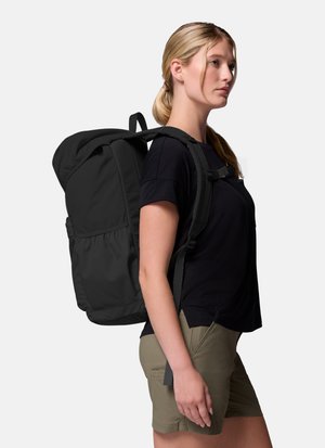Kobieta o blond włosach, ubrana w czarną koszulkę i khaki krótkie spodenki, niesie duży czarny plecak z wyściełanymi paskami na ramiona i klamrą na klatce piersiowej.