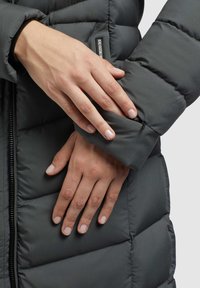 Doudoune gris foncé avec texture matelassée, dotée d'une poche zippée sur la manche et d'un col montant. Tissu lisse et mat.