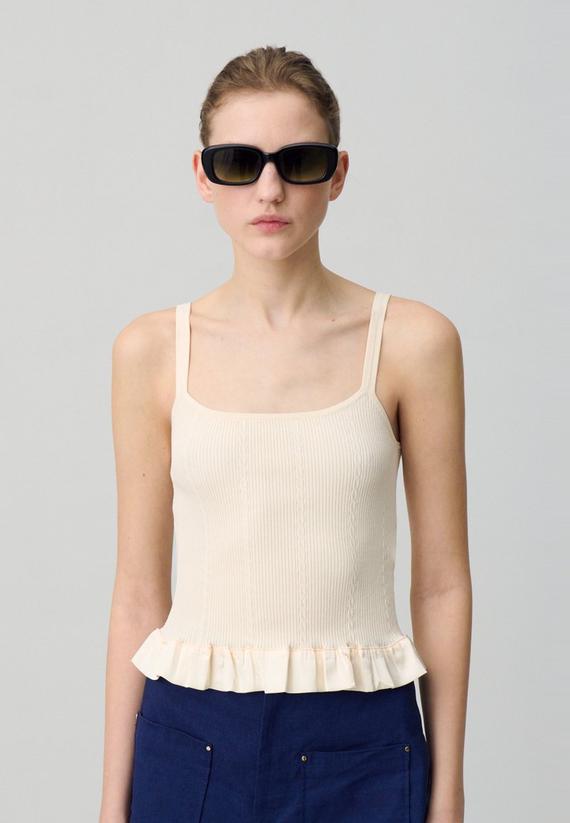 Débardeur beige à rayures avec des bretelles fines, doté d'un ourlet à volants. Associé à un bas marine. Modèle portant des lunettes de soleil noires.